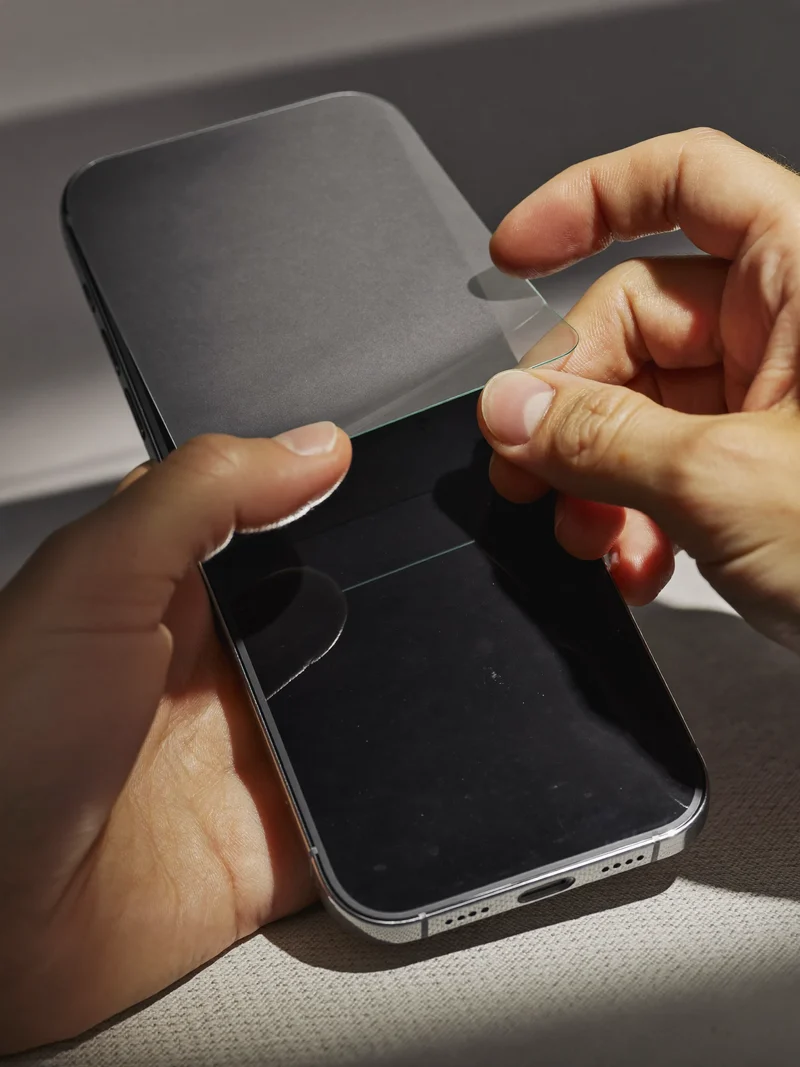 Detailní záběr rukou při lepení tvrzeného skla na telefon s prázdným displejem.